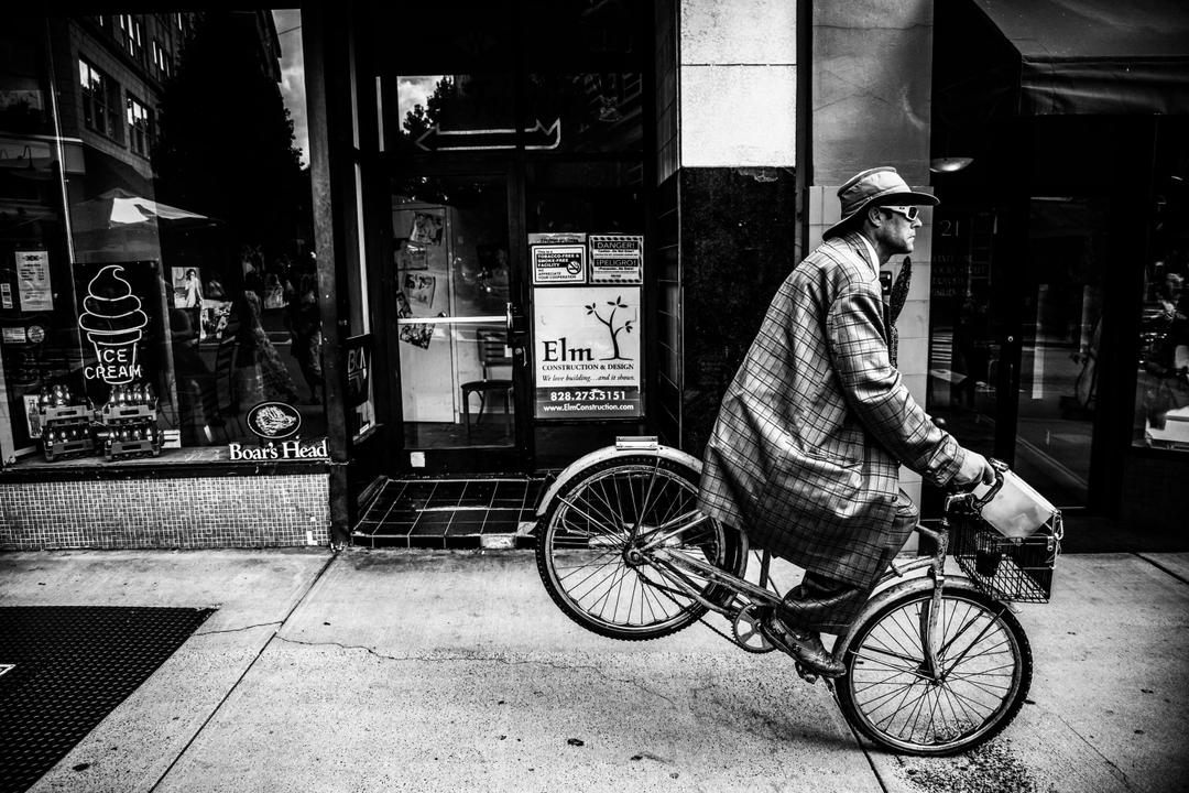 Photograph of a sharply dressed man with cheap sunglasses doing a stoppie (front wheelie) on a bicycle in front of a shop in downtown Huntsville, AL 