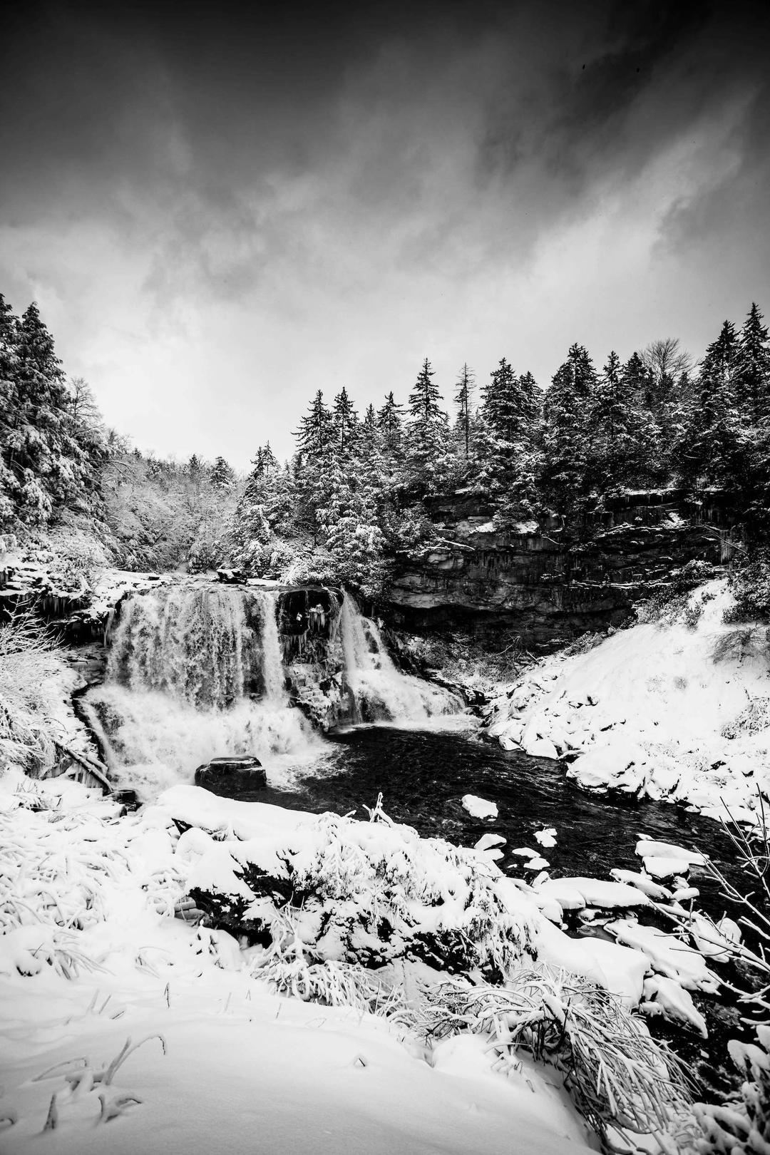 Black and white photograph of snowcapped woods surrounding a waterfall and river